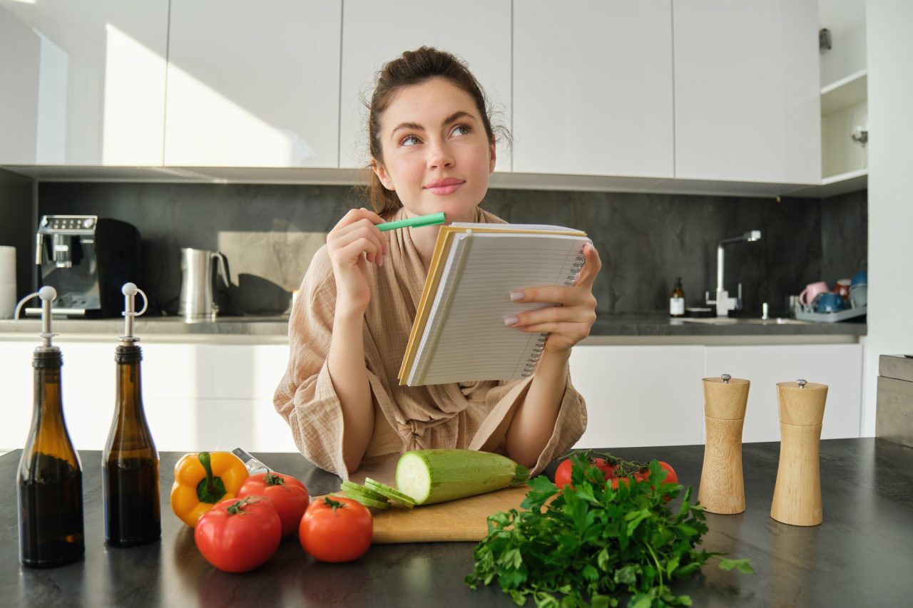 portrait of woman checking recipe notes in notebook standing in kitchen with vegetables cooking